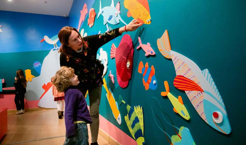mamá junto con su hijo al interior de la sala, ella esta señalando un muro donde hay peces de colores ilustrados con relieves en el muro.